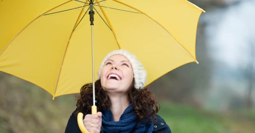 Femeie cu părul creț și brunet, râzând cu bucurie sub un umbrelă galbenă, purtând o căciulă albă și o eșarfă albastră
