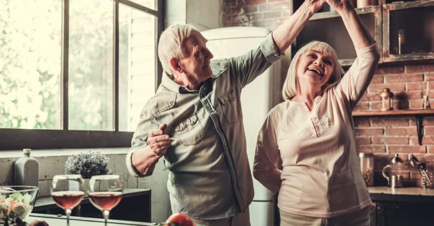 Un cuplu în vârstă dansează și râde împreună într-o bucătărie, cu un bărbat îmbrăcat într-o cămașă kaki și o femeie într-un top crem.